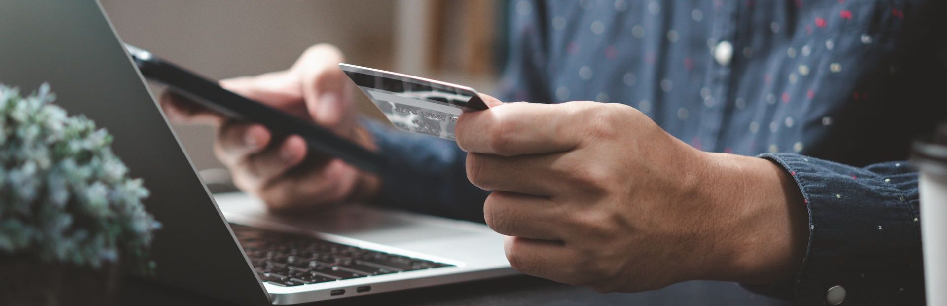 Vyriškos rankos prie nešiojamojo kompiuterio, viena ranka laiko telefoną, kita – mokėjimo kortelę.